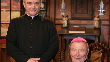 Pfarrer Anton Pfaffinger (Götz Burger, links) freut sich über den Besuch des Bischofs (Erich Hallhuber sen.). | Bild: BR/Foto Sessner