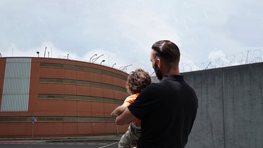 Ein Mann mit Kind auf dem Arm steht vor einem Gefängnis.  | Bild: BR/ Carina Müller 
