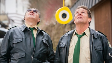 Hubert (Christian Tramitz, rechts) und Reimund Girwidz (Michael Brandner) sehen nach oben, wo Staller vom Brückengeländer den Abstand zum Boden misst. | Bild: ARD/TMG/Arvid Uhlig