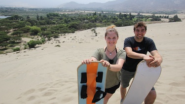 Trujillo, Peru: Ausflug der Praktikanten Günther Schön und Laura Bauer zum Sandborden. Die beiden Praktikanten sind schon früh auf den Beinen. Sie wollen heute Sandborden ausprobieren. Bei Lehrer Vicent und Übersetzerin Aurelia dürfen sie Bords aussuchen. Etwa 15 Minuten laufen sie zur Düne, wo sie sich erst einmal aufwärmen müssen. Anschließend geht es die Düne hinauf, doch Laura hat nur Birkenstocks an und verbrennt sich die Füße. Sie kann nicht mehr weiterlaufen. Also leiht Günther ihr seine Schuhe und geht mit Socken die Dünen rauf. Oben angekommen zeigt Guide Vicent, dass sie im Stehen und im Liegen runterfahren können. Die beiden fahren im Stehen. | Bild: BR/Bewegte Zeiten Filmproduktion GmbH/André Goerschel