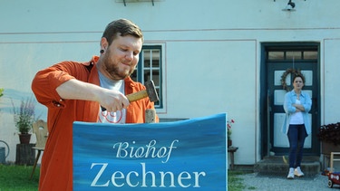 Severin (Andi Gießer) hämmert stolz das neue Bio-Hof Schild in den Boden. | Bild: BR/Marco Orlando Pichler