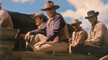 Die Söhne der Katie Elder – John (John Wayne, zweiter von links), der älteste und ein Revolverheld; Tom (Dean Martin, rechts), ein professioneller Spieler; Matt (Earl Holliman, zweiter von rechts), ein erfolgloser Eisenwarenhändler; und der jüngste, Bud (Michael Anderson Jr., links) – waren jahrelang nicht mehr zu Haus und sind zur Beerdigung angereist. Doch die Bewohner von Clearwater stehen ihnen recht ablehnend gegenüber, da sie sich nicht sehr um ihre Mutter gekümmert hatten. | Bild: BR/Paramount Pictures