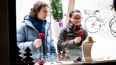 Lien (Nui Nguyen) steht mit Till (Marinus Hohmann) vor einem Schaufenster und versucht subtil ihm einen Tipp zu geben, was ihr gefällt. | Bild: BR/Lena Semmelroggen