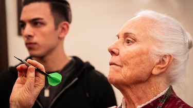 Theresa (Ursula Erber) spielt Darts. Im Hintergrund: Leon (Joel Akgün). | Bild: BR/Lena Semmelroggen