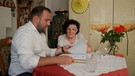 Ein Mann und eine Frau sitzen an einem Tisch in einer Wohnung und sichten Akten | Bild: BR
