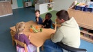 Drei Kindergartenkinder und ein Erwachsener sitzen um Tischchen und essen Gemüse | Bild: BR