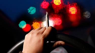 Cannabis und Autofahren - Symbolbild: Ein Joint wird am Lenkrad gehalten. | Bild: picture alliance / Panama Pictures | Christoph Hardt