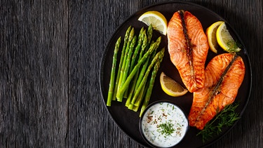 Zwei gegrillte Lachssteakscheiben mit grünem Spargel, Joghurt-Dillsauce und frischen Zitronenscheiben auf einem großen schwarzen Teller angeordnet. | Bild: stock.adobe.com/myviewpoint