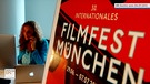 Links Diana Iljine vor einem Computer sitzend, telefonierend, rechts Plakat zum 30. Filmfest München | Bild: BR Archiv