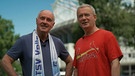 Zwei Männer in T-Shirts, einer davon mit eiem Fan-Schal, posieren vor einem Sportstadion. | Bild: BR