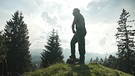 Ein Mann in Arbeitskleidung steht auf einer Almwiese mit Nadelbäumen und blickt in das Bergpanorama. | Bild: BR