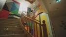 Eine farbenfrohe Treppe innerhalb der Villa K. An der Wand, entlang der Treppe, sind viele unterschiedliche Farben und erinnert an die Kunstrichtung ArtPop. | Bild: BR