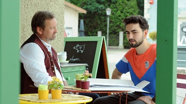 Dahom is Dahoam: Gregor (Holger Matthias Wilhelm, links) sitzt mit Felix (Shayan Hartmann, rechts) im Schanigarten. | Bild: BR/Marco Orlando Pichler