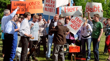 Ganz Lansing demonstriert gegen Gerstl vom Landratsamt Baierkofen, damit Xaver weiter auf seiner Wiese wohnen darf. | Bild: BR/Marco Orlando Pichler