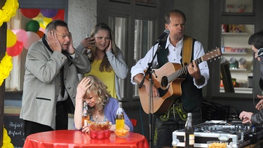 Die Eröffnungsfeier von Kiosk und Jugendtreff verläuft äußerst skurril. Von links: Alois Preissinger (Erich Hallhuber sen.), Caro Ertl (Teresa Rizos), Yvonne Preissinger (Alexa Eilers) und ein Schlagersänger (Andreas Lipperer). | Bild: BR/Marco Orlando Pichler