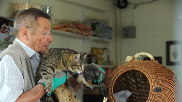 Damit der Kater Schatzi den Kiosk nicht weiter durcheinander bringt, sperrt Alois (Erich Hallhuber sen.) ihn wieder in seinen Käfig ein. | Bild: BR/Marco Orlando Pichler