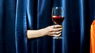 Ein Glas Rotwein wird von einer Frauenhand durch einen blauen Vorhang gereicht. | Bild: BR/ Monika Rasciute, Hannah Wiesner