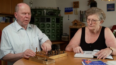 Ludwig Zier sitzt vor seinem Blinden-Schachspiel, neben ihm blätterst seine Lebensgefährtin in einem Buch. | Bild: BR
