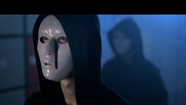 Eine Person mit weißer Maske und schwarzer Kapuze und einer roten Träne aus dem Musikvideo zu "Tausend Farben". | Bild: BR