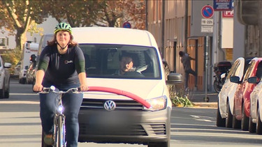 Fahrradfahrerin mit Poolnudel auf dem Sattel, fährt vor einem PKW auf der Straße. | Bild: BR