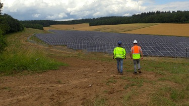 Der Solarpark in Fuchsstadt mit zwei Mitarbeitern die davor stehen. | Bild: BR