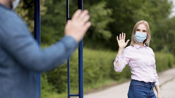 Frau mit Maske begrüßt mit Abstand ihr Gegenüber. In der Corona Pandemie galten Abstandsregeln bei sozialen Begegnungen.  | Bild: picture-alliance/ imageBROKER  Oleksandr Latkun