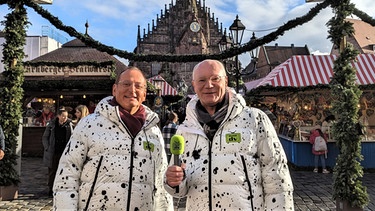 Stefan Strohstern (Martin Rassau, rechts) und Theo Dolores Hanfgruber (Volker Heißmann) vom fränkischen Sender"Doldi-DV" stürzen sich getreu ihrem Motto "Mitten drin und voll daneben" in das bunte Christkindlesmarkt-Getümmel.  | Bild: BR/Amelie Völker