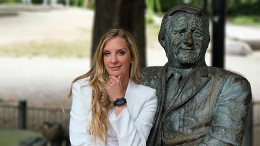 Moderatorin Caro Matzko am Denkmal für Helmut Fischer als Monaco Franze vor dem Café "Münchner Freiheit". | Bild: BR/Björn Pfeffermann