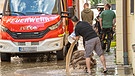 Ein Mann schippt schüttet einen Eimer Wasser neben Sandsäcken auf die überschwemmte Strasse. Im Hintergrund ein Feuerwehrauto und Einsatzkräfte. | Bild: picture alliance / Wolfgang Maria Weber | R7172