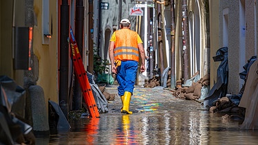 Ein Helfer in Gummistiefeln geht durch eine überflutete Strasse. | Bild: picture alliance/dpa | Armin Weigel