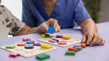 Eine Altenpflegerin spielt mit einer Rentnerin mit einem Puzzle aus Holz. | Bild: stock.adobe.com/toa555