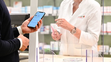 ARCHIV - 29.09.2021, Niedersachsen, Oldenburg: Ein Mann mit seinem Handy in der Hand, auf dem die App «Das E-Rezept» geöffnet ist, wird in einer Apotheke von einer Apothekerin bedient (zu dpa Bundestag soll Schub für E-Rezept und E-Patientenakte beschließen»). Foto: Mohssen Assanimoghaddam/dpa +++ dpa-Bildfunk +++ | Bild: dpa-Bildfunk/Mohssen Assanimoghaddam