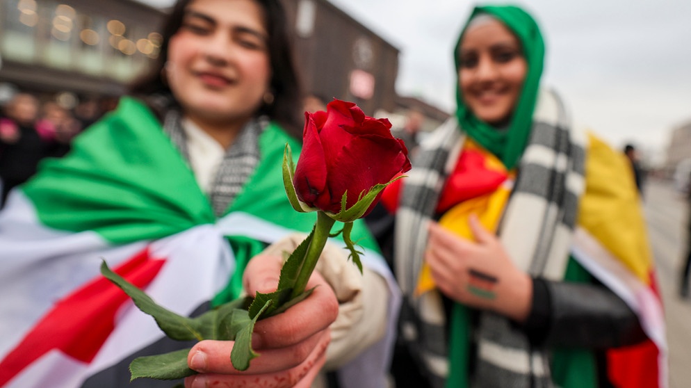 10.12.2024, Nordrhein-Westfalen, Duisburg: Zwei Frauen zeigen eine rote Rose zum Zeichen des Friedens in Syrien, tragen die Flagge der syrischen Revolution (l) und die deutsche Flagge um die Schultern als sie vor dem Hauptbahnhog Duisburg feiert. Feiernde hatten Flaggen der syrischen Revolution gezeigt und gejubelt. Die Flagge der Gegner des Assad-Regimes zeigt drei horizontale Streifen - einen grünen, einen weißen und einen schwarzen - sowie drei rote Sterne. Foto: Christoph Reichwein/dpa +++ dpa-Bildfunk +++ | Bild: dpa-Bildfunk/Christoph Reichwein