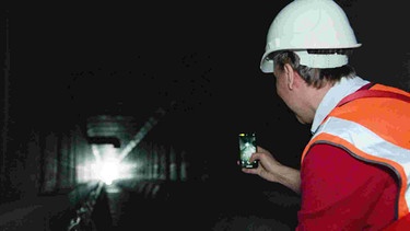 Mann in Schutzkleidung fotografiert mit seinem Handy Wärmetauschermodule in einem Abwasserkanal. Ansicht von hinten.
| Bild: BR