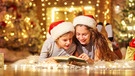 Zwei Kinder lesen vor einem Weihnachtsbaum ein Buch | Bild: Colourbox