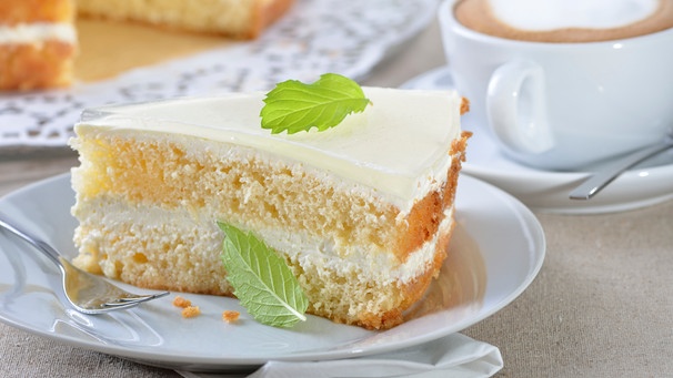 Ein Stück Joghurttorte auf einem weißen Kuchenteller mit einer Tasse Cappucchino. | Bild: mauritius images / Pitopia / Karl Allgäuer