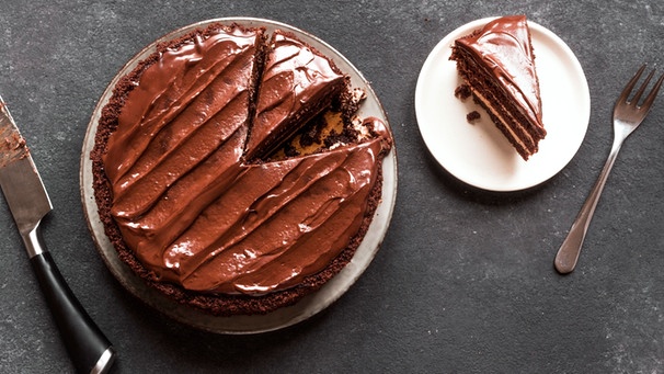 Eine angeschnittene Schokoladentorte mit einer glänzenden Schokoladenganache auf einer grauen Schieferplatte. Neben der Torte ist ein Stück der Torte auf einem weißen Kuchenteller. | Bild: mauritius images / Oksana Bratanova / Alamy