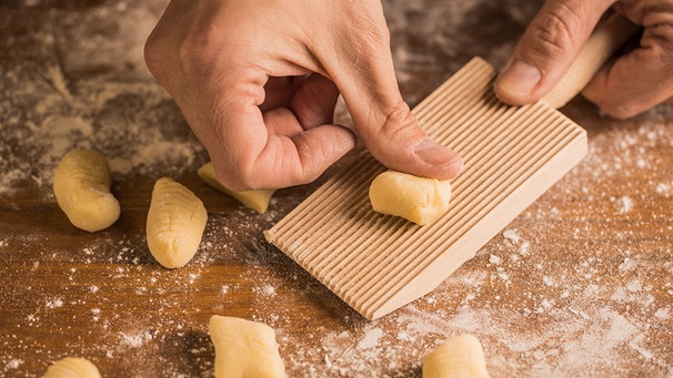 Kochhände drücken ein Stück Teig auf ein geripptes Brett, während sie Gnocchi auf einem mit Mehl bedeckten Holztisch zubereiten; | Bild: mauritius images / The Picture Pantry / Ramón López Farinós