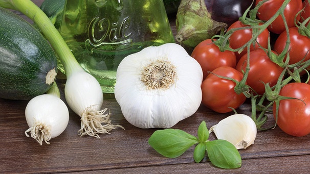 Nahansicht einer Zucchini, Auberginen, einer Knoblauchknolle , Zwiebeln, Basilikum und Tomaten auf einem Holztisch. Eine Flasche mit der Aufschrift "Öl" steht im Hintergrund. | Bild: picture alliance / Panther Media | Jaroslav Frank