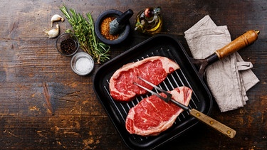 Eine Grillpfanne von oben fotografiert mit zwei Steaks und einer Fleischgabel. Die Pfanne steht auf einem braunen Holztisch, unter der Pfanne ist ein weißer Lappen. Neben der Pfanne sind ein Schälchen mit einem Mörser und Gewürzen. | Bild: mauritius images / TPP / Natalia Lisovskaya