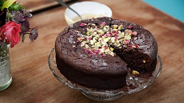 Ein runder angeschnittener Schokoladenkuchen mit Pistazien und Rosenblättern auf einer Glasplatte, die auf einem braunen Holztisch steht. Daneben ist ein Blumenstrauß in einer Glasvase angeschnitten zu sehen und hinter dem Kuchen ein Schälchen mit einem kleinen Löffel darin. | Bild: mauritius images / foodcollection / Steven Joyce