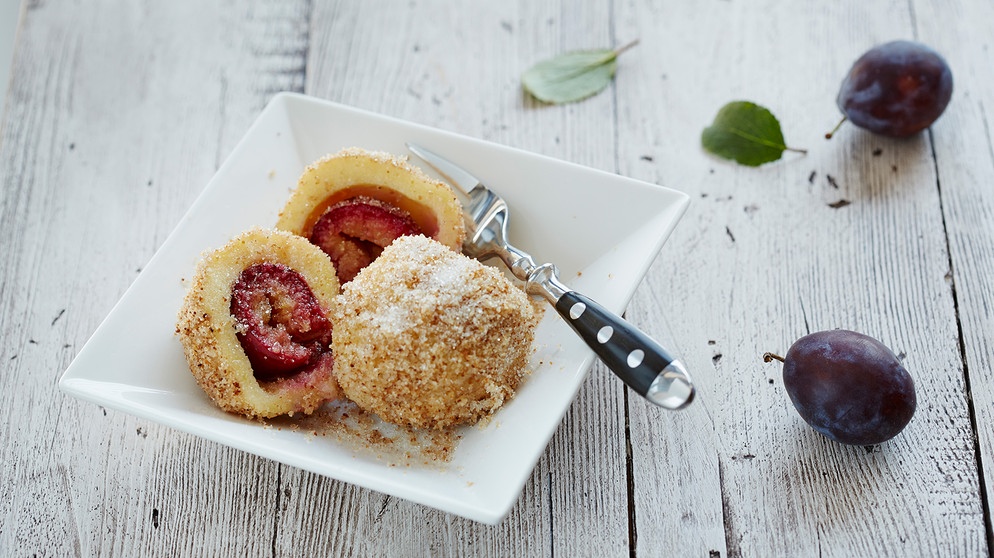 ein ganzer und ein durchgeschnittener Zwetschgenknödel auf einem weißen Teller mit einer Gabel auf einem hellen Holztisch | Bild: mauritius images / Westend61 / Daniel Schweinert