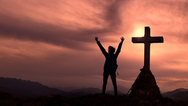 Kreuz auf dem Gipfel des Berges mit einem Bergsteiger, der die Hände hebt | Bild: picture alliance / CHROMORANGE / Michelangelo Oprandi