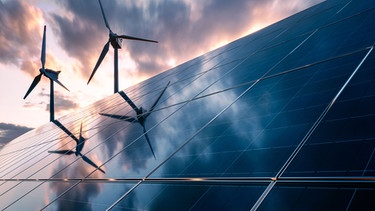 Woher kommt die Sonnenenergie? - Eine Solaranlage auf einem Feld mit Windrädern im Hintergrund. | Bild: stock.adobe.com/PhotoGranary