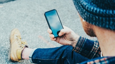 Ein junger Mann sitzt am Straßenrand und bedient sein Smartphone. | Bild: stock.adobe.com/Alex from the Rock