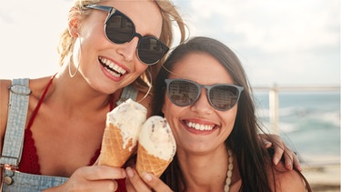 Zwei junge Frauen halten lachend ein Eis in der Hand. Welches Eis hat die wenigsten Kalorien? Welche Eissorte essen wir am liebsten? Und wer hat das Speiseeis eigentlich erfunden? Wir haben 6 Fakten über Eis für euch.  | Bild: Wochit/Getty Images