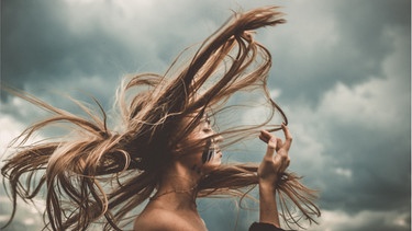 Portrait einer jungen Frau mit wehenden hellbraunen Haaren vor wolkigem Hintergrund. Spliss kann man reparieren, Stress kann zu Haarausfall führen und durch regelmäßiges Spitzenschneiden wachsen Haare schneller. Sind diese Haar-Mythen wahr oder falsch?  | Bild: Wochit/Getty Images