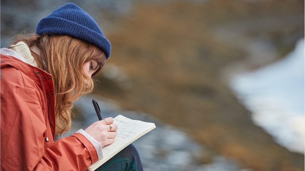 Eine junge Frau schreibt Tagebuch an einem See. Wir erklären, warum euch Tagebuchschreiben auch im Erwachsenenalter viel bringen kann und haben Tipps, die euch beim Schreiben helfen. | Bild: Wochit/Getty Images