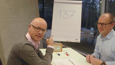 Karsten Schwanke und Professor Christian Hesse sitzen an einem Tisch, im Hintergrund ist ein Whiteboard mit der Zahl 137. | Bild: BR