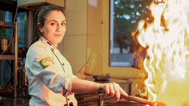 Eine junge Frau steht schmunzelnd vor einem Grillherd mit offener Flamme. | Bild: hr/isar film Produktion GmbH